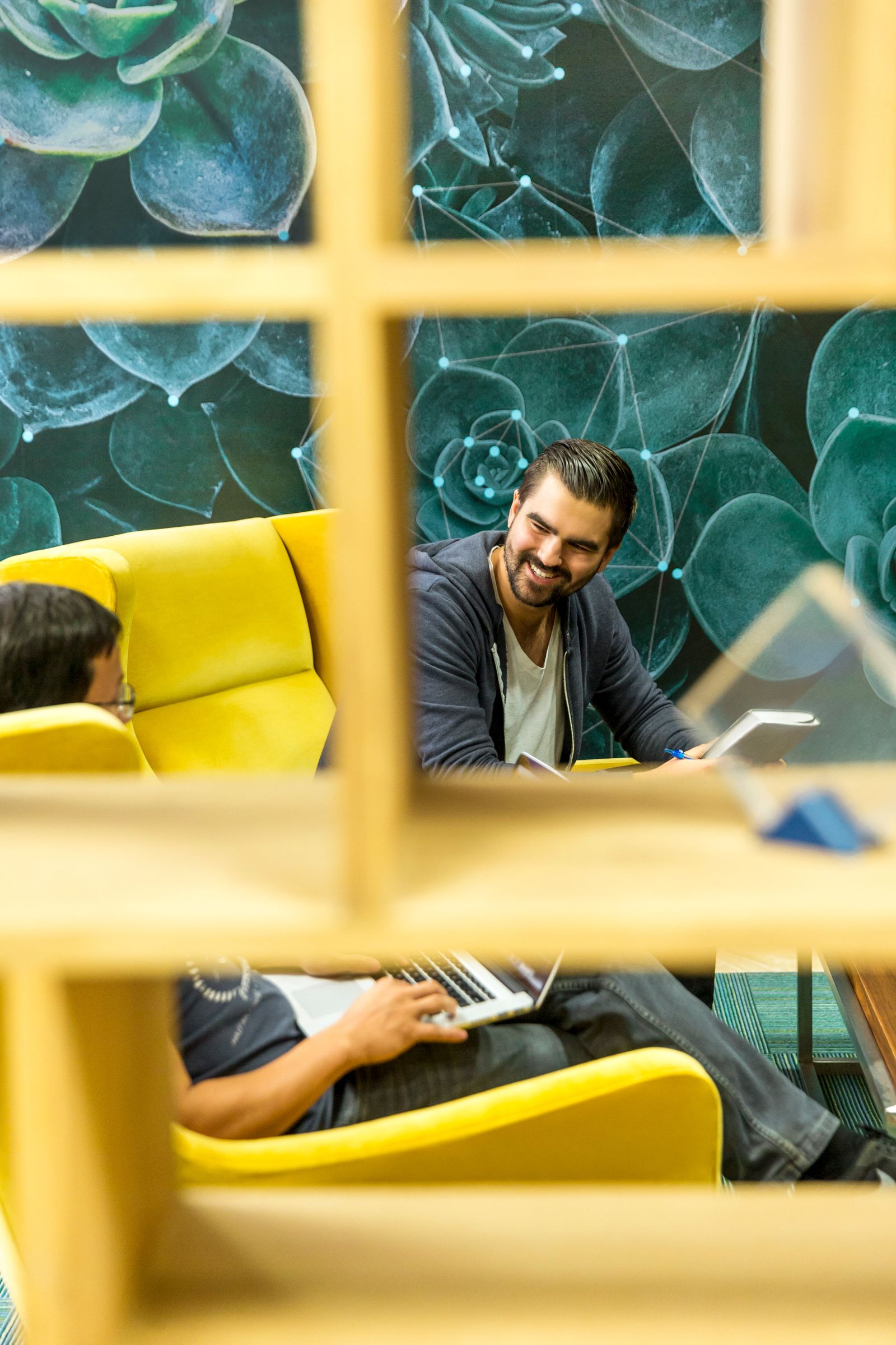 Zwei Männer mit Laptops und Notizbüchern sitzen auf gelben Sesseln in einer Lounge, gesehen durch ein Holzregal.
