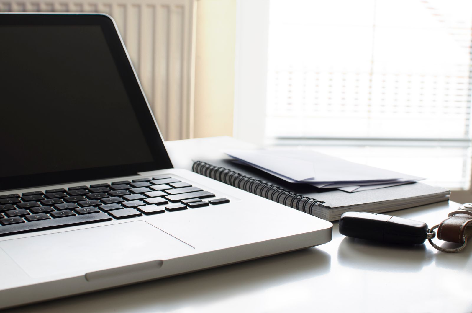 Ein offener Laptop, ein Spiralnotizbuch mit Briefumschlägen und ein Autoschlüssel auf einem weissen Schreibtisch am Fenster.